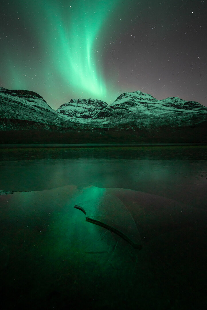 Bright Aurora reflects its light over a frozen lake that is in the foreground of some mountains