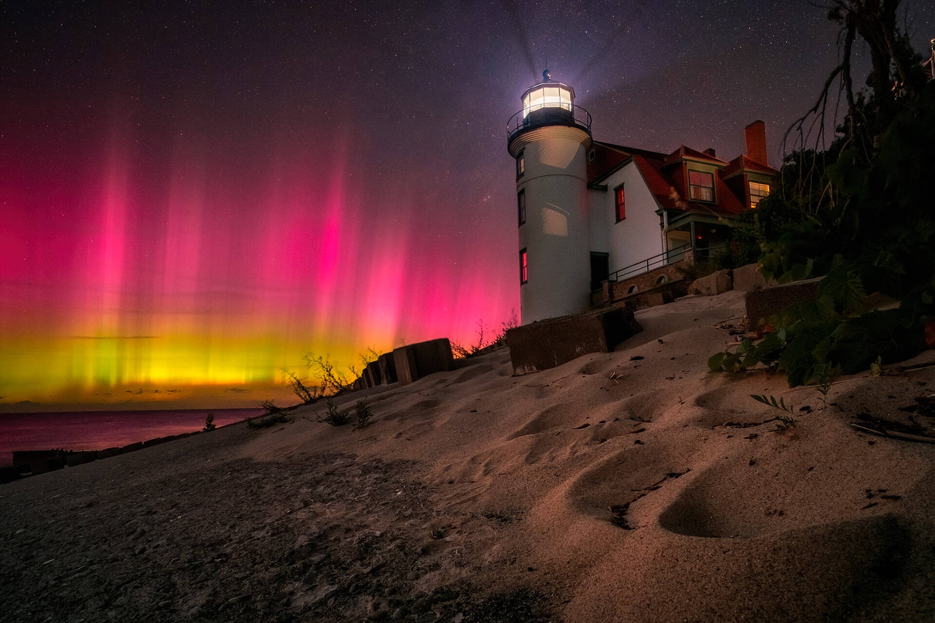 Aurora Boreal de color rojo intenso y brillante cubre el cielo nocturno tras un faro en una playa