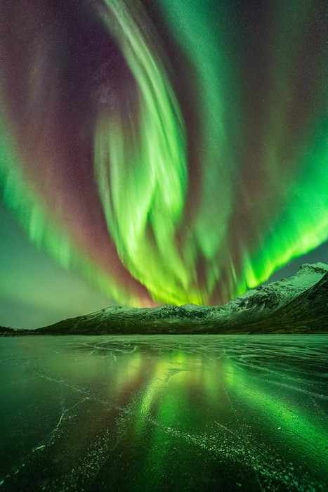 Northern lights covering the sky with bright green and red colors and reflecting light over frozen lake