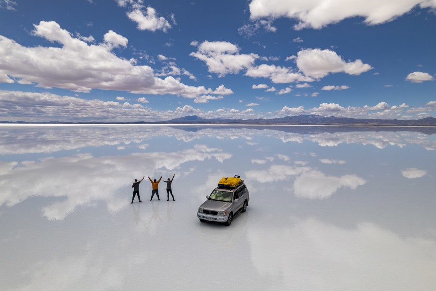 Cloudy sky reflected in Salar de Uyuni with an SUV and three people
