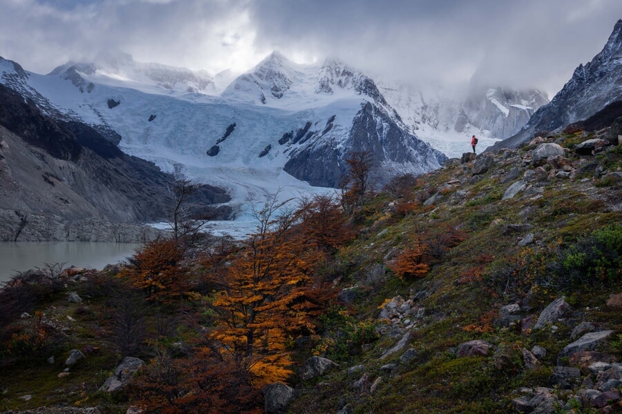 A man stands atop a rocky hill, admiring the view of a lake and mountains in the distance